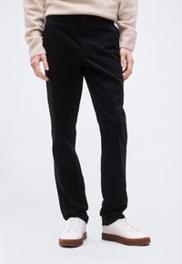 Άτομο που φοράει μαύρο παντελόνι ίσια γραμμή με λευκά αθλητικά παπούτσια και μπεζ πουλόβερ, όρθιο μπροστά σε απλό ανοιχτό φόντο.