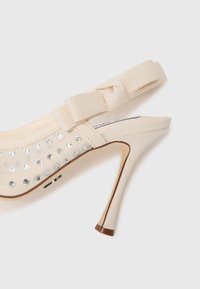 Beige high-heeled slingback shoe with rhinestone accents, fabric upper, wide elastic strap, and a pointed toe. Slightly curved heel with wooden detail.