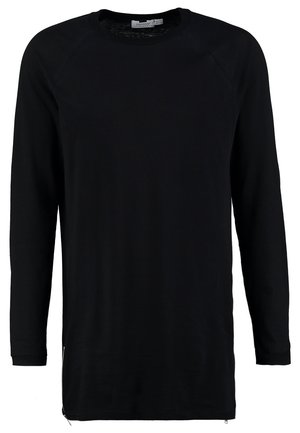 Homme portant une veste à capuche noire, une chemise noire, un jean gris et des bottines noires, debout devant un fond clair uni.