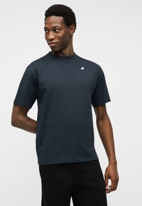 Νέος άνδρας που φοράει σκούρο μπλε κοντομάνικο μπλουζάκι με μικρό λευκό λογότυπο, κοιτάζοντας προς τα αριστερά του, με απλό φωτεινό φόντο.