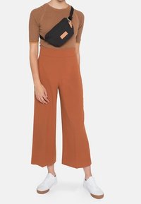 Une personne portant un haut marron à manches courtes, un pantalon large taille haute rouille, des baskets blanches et un sac bandoulière noir sur un fond clair.