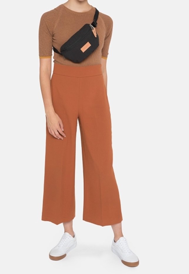 Une personne portant un haut marron à manches courtes, un pantalon large taille haute rouille, des baskets blanches et un sac bandoulière noir sur un fond clair.