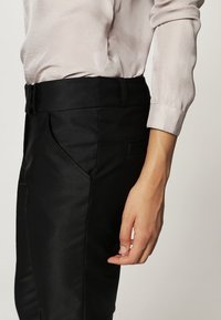 Svarta skräddarsydda byxor med en slät textur, med sidfickor och ett något avsmalnat ben, i kombination med en ljusfärgad blus.