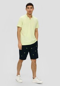 Hellgrünes Polohemd mit V-Ausschnitt, kombiniert mit dunkelblauen Shorts mit grünen Blätternmuster und weißen Turnschuhen. Modell steht frontal.