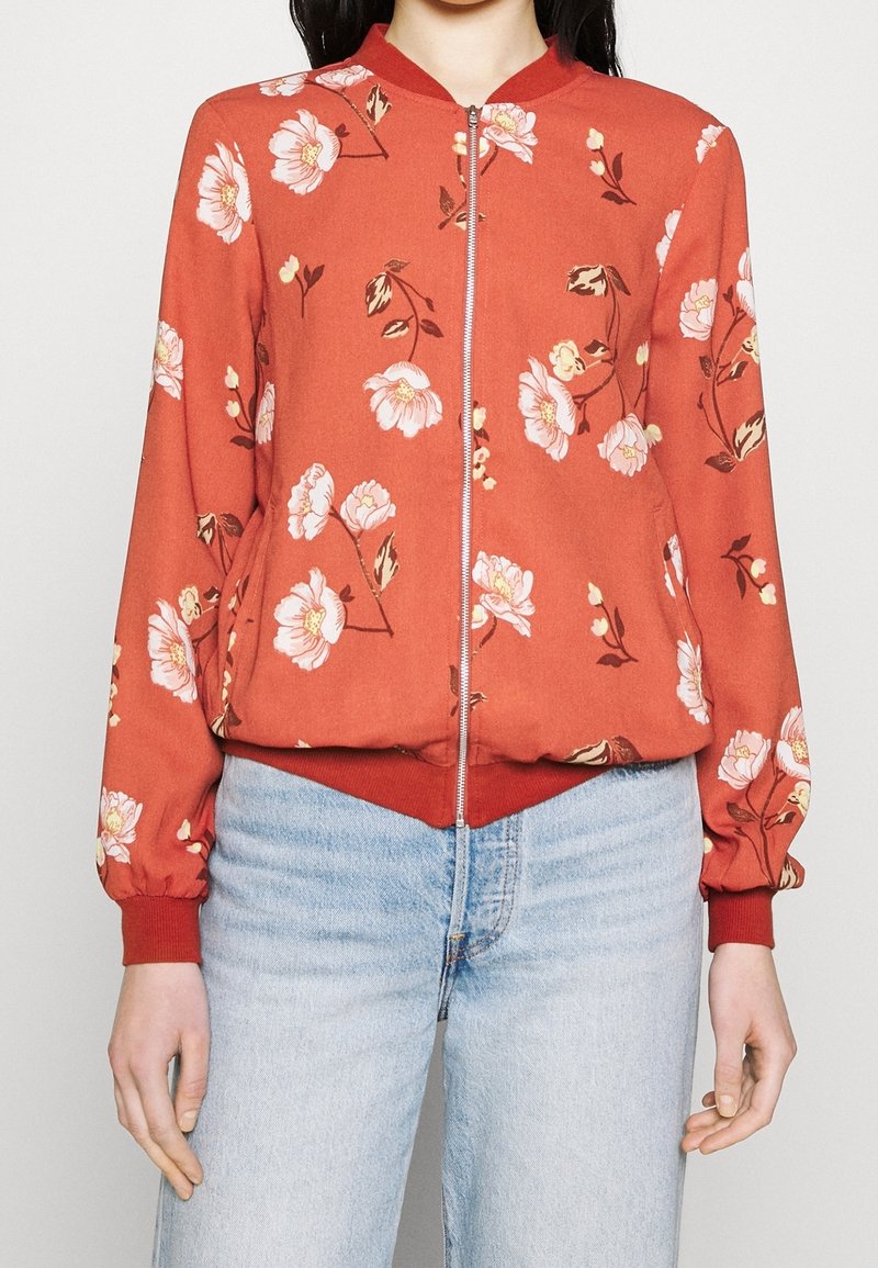 Veste bomber rouge à fleurs avec fermeture éclair sur le devant, poignets et ourlet côtelés ; présente des motifs floraux surdimensionnés en rose et blanc sur un tissu doux.