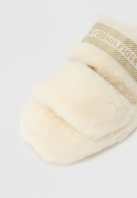 Pluizige crèmekleurige pantoffel met drie gevoerde banden, voorzien van een textuur van beige merkband. Zacht materiaal voor comfort.
