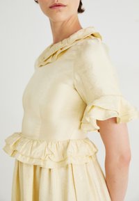 Gaiša dzeltena kleita ar ruffle apkakli un piedurknēm, ar pakāpeniskām ruffles malām, izgatavota no mīksta, gluda auduma.