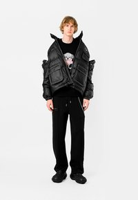 Chaqueta acolchada negra con grandes bolsillos, cierre de cremallera y puños elásticos. Llevada sobre un suéter negro con un gráfico colorido de un oso y combinada con pantalones negros y zapatos texturizados.