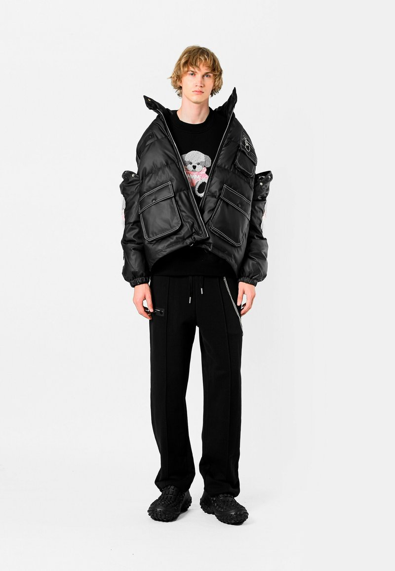 Chaqueta acolchada negra con grandes bolsillos, cierre de cremallera y puños elásticos. Llevada sobre un suéter negro con un gráfico colorido de un oso y combinada con pantalones negros y zapatos texturizados.