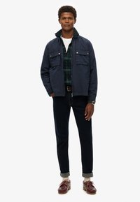 Navy blue jacket with two chest pockets over a green plaid shirt, paired with dark jeans and burgundy shoes. Casual outfit with mixed textures.