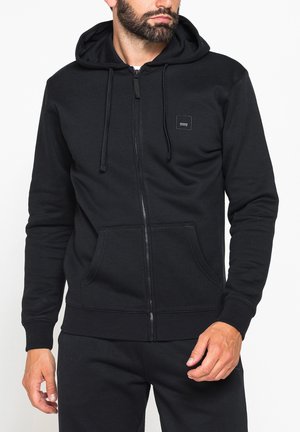 Uomo con felpa nera con cappuccio e cerniera, tasche frontali a marsupio e coulisse, abbinata a pantaloni neri su sfondo bianco.