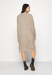 Vestito maglione beige a maglia con maniche lunghe, lunghezza ginocchio, scollo rotondo e aperture laterali. Abbinato a stivali neri alla caviglia con dettagli dorati.
