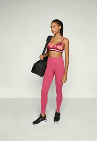 Junge Frau in rosa Sportbekleidung, die eine schwarze Sporttasche über der Schulter hält, steht vor einem schlichten, hellen Hintergrund.