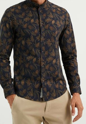 Homme portant une chemise noire slim à manches longues avec un motif de feuilles marron, boutonnée devant, et un pantalon beige, les mains partiellement dans les poches.