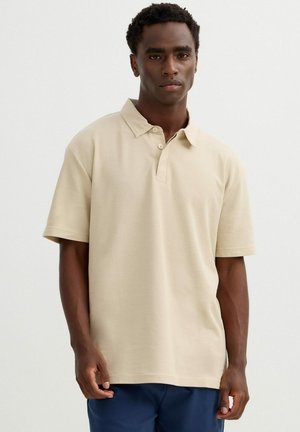 Hombre vistiendo una camiseta tipo polo beige de manga corta y pantalones azul marino contra un fondo claro y simple.