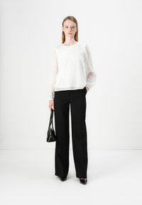 White textured long-sleeve blouse with wavy pattern, paired with black high-waisted trousers and pointed black heels. Model carries small black bag.