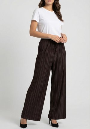 Femme portant un haut blanc à manches courtes, un pantalon marron plissé à jambes larges et des chaussures noires à talons hauts, debout devant un fond uni.