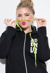 Blonde woman with a top bun, wearing a black zip-up hoodie, holds neon yellow drawstrings stretched under her nose, puckering lips playfully.