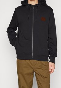 Felpa nera con zip in tessuto morbido e elasticizzato; presenta una tasca frontale, cappuccio con cordini e un'etichetta in tessuto marrone testurizzato sul petto.