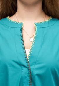 Γυναίκα που φοράει τοπ σε τιρκουάζ χρώμα με κίτρινη και καφέ διακοσμητική ραφή, λευκό μπλουζάκι από μέσα και χρυσή αλυσίδα με στρογγυλό μενταγιόν.