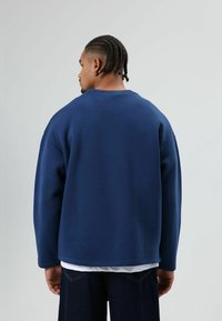 Marineblauwe, oversized sweatshirt met een ronde hals, lange mouwen en een gladde textuur, gedragen met een wit overhemd eronder en donkere spijkerbroek.