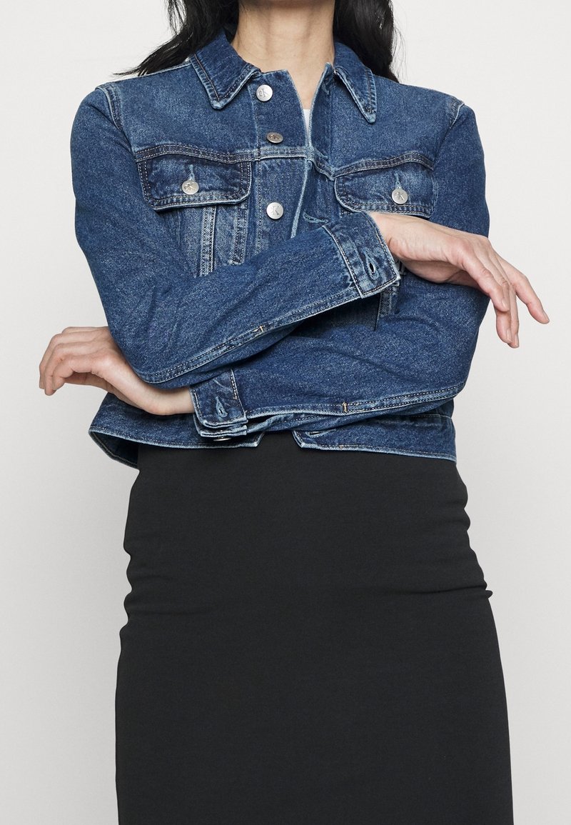 Kraftigt denimjacka i mörkblått, med två framfickor, silverknappar och klassisk krage. Buren över en svart kjol.