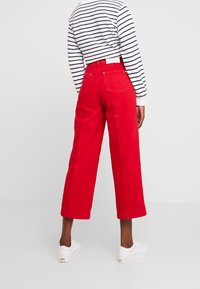 Persona che indossa pantaloni a gamba larga rossi, scarpe da tennis bianche e una maglia a maniche lunghe bianca con righe orizzontali nere, di spalle.