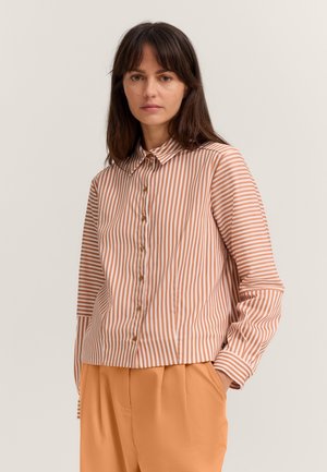 Frau trägt ein beige-weiß gestreiftes Hemd mit Knöpfen und hellbraune Hose, steht mit den Händen in den Taschen vor einfachem Hintergrund.