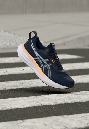 Marineblauer Laufschuh mit weißer Sohle und pastellorangefarbenem Akzent, in der Luft über einem schwarz-weiß gestreiften Zebrastreifen.