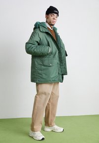 Zelená prošívaná bunda s texturovanou kapucí, předními kapsami a kovovými patentními knoflíky, doplněná o hnědé kalhoty a bílé tenisky.