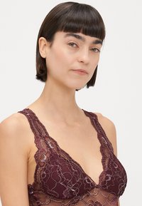 Čipkasti bralet u burgundcu s cvjetnim vezom, V-izrezom i valovitim rubovima. Mekana, prozračna tekstura s složenim detaljima.