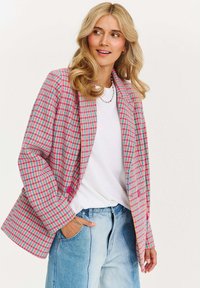 Žena se blond vlasy, oblečená v růžovo-modrém károvaném blejzru přes bílou košili a světle modré džíny, kouká do strany s rukou v kapse.