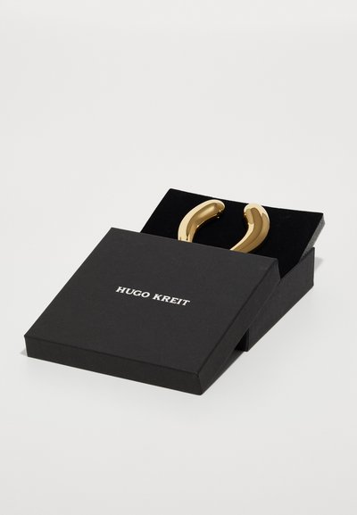Pendientes de metal en tono dorado con un diseño suave y curvado, presentados en una caja negra con un logo blanco en la parte superior.