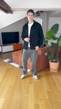 Moški v črni jakni, beli srajci, sivih hlačah in športnih copatih stoji v dnevni sobi s tremi elementi: rjavim kavčem, lesenim podom, zeleno rastlino in televizorjem.