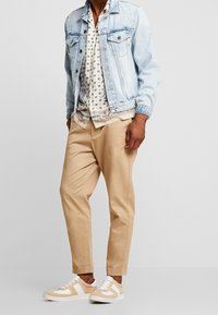 Mies joka käyttää vaaleansinistä denim-takkia, kuviollista nappipaitaa, beigejä kapeita housuja ja beige-valkoisia lenkkareita seisoo tasaisella taustalla.