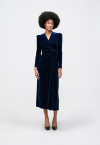 Ženska z afro pričesko nosi dolg rokav obleko iz modrega žameta in črne visoke pete, stoji ob svetli enobarvni ozadju.