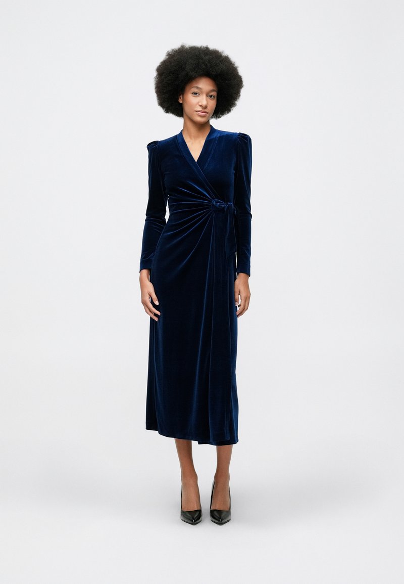 Ženska z afro pričesko nosi dolg rokav obleko iz modrega žameta in črne visoke pete, stoji ob svetli enobarvni ozadju.