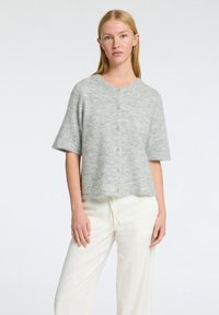 Cardigan corto grigio chiaro a maniche corte, vestibilità ampia, lavorazione a maglia testurizzata, chiusura con cinque bottoni, scollatura rotonda, abbinato a pantaloni a gamba larga color crema.