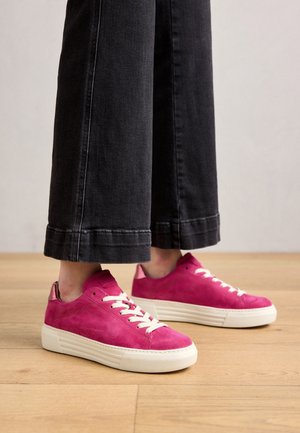 Stopy w jasno różowych zamszowych trampkach z białymi sznurówkami i podeszwami, w połączeniu z czarnymi, krótkimi spodniami o szerokich nogawkach na drewnianej podłodze.