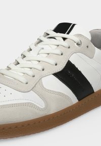 Baskets blanches et beiges avec des lacets blancs, une bande noire sur le côté, et une semelle en caoutchouc marron sur un fond uni.