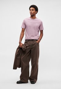 Νέος άνδρας που φοράει ένα ανοιχτό μωβ μπλουζάκι και φαρδιά καφέ παντελόνια, κρατώντας ένα καφέ σακάκι στο αριστερό του χέρι, στέκεται μπροστά σε απλό φόντο.