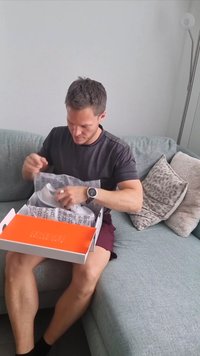 Biały pudełko z pomarańczowym wnętrzem, wypełnione zmiętą bibułą. Osoba rozpakowuje je, siedząc na fakturowanej, jasnoniebieskiej kanapie.