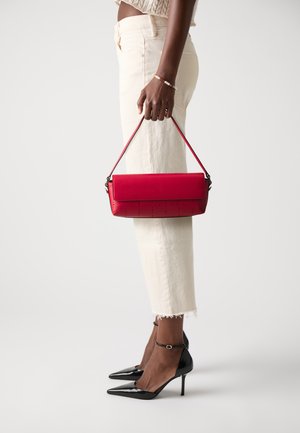 Borsa a spalla in pelle rossa con patta, dettagli perforati e tracolla corta, tenuta da una persona che indossa pantaloni cream a vita alta e tacchi neri.
