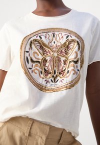 Bílý bavlněný tričko s vyšitým motivem motýla v zlaté, růžové a černé barvě, rámované dekorativním okrajem se svátečními vzory.