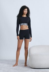 Must mustriga pusa "DIESEL" logoga, mille juurde on kombineeritud mustad lühikesed püksid. Modell seisab paljajalu heledat värvi vaiba peal, kõrval on beež kõverdiivani.