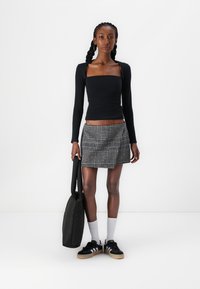 Zwarte langslavige top met vierkante halslijn, grijs en zwarte houndstooth geplooide rok, zwarte sneakers, witte sokken en een zwarte draagtas.