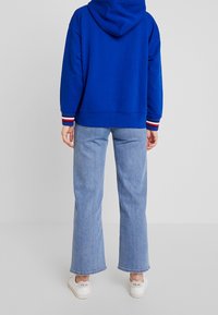 Modrá mikina s kapucí a pruhovanými manžetami. Kombinována s lehce modrými kalhotami se širokými nohavicemi a bílými teniskami. Jednobarevné pozadí.