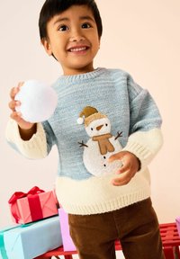 Pull en tricot bleu clair avec un motif de bonhomme de neige, poignets couleur crème et pantalons en velours côtelé marron. Tenant une boule de neige blanche et moelleuse.