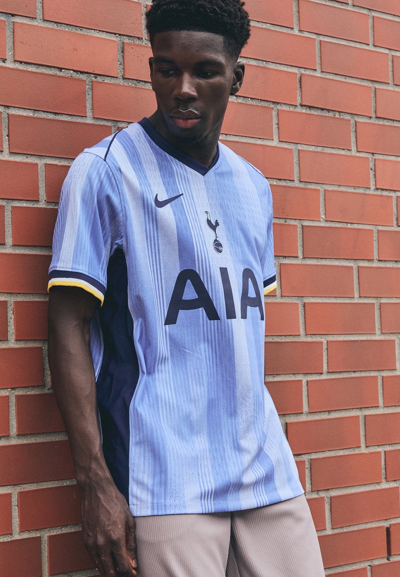 Jasnoniebieska koszulka piłkarska z pionowymi paskami, logo AIA w wyraźnej czerni oraz żółtymi akcentami na rękawach. Logo Nike wyraźnie widoczne.