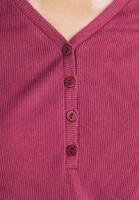 Ribbade burgunderfärgad topp med V-ringning, prydd med fyra matchande knappar längs mittfram. Tyget har en texturerad yta.